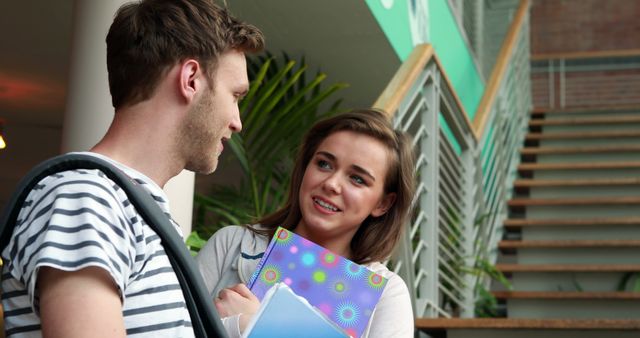 College students having a casual conversation in a staircase area on campus. Both students appear engaged and happy, holding books while chatting informally. This image is ideal for use in educational materials, marketing for universities and colleges, or articles about student life, interactions, and campus atmosphere.