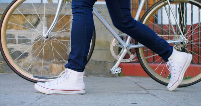Person wearing casual jeans and white sneakers walking alongside a vintage bicycle on an urban street. Suitable for use in transportation, urban lifestyle, or fitness-related projects. This image can enhance articles or blogs focusing on city commuting, personal transportation choices, or healthy urban lifestyles.