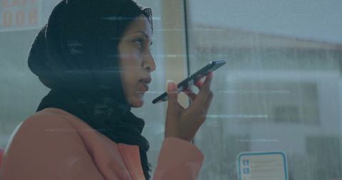 Muslim woman wearing hijab speaking into smartphone on bus during urban commute, candid