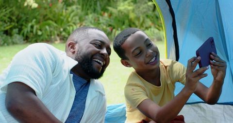 African american father and son sharing smartphone in backyard tent, laughing and bonding