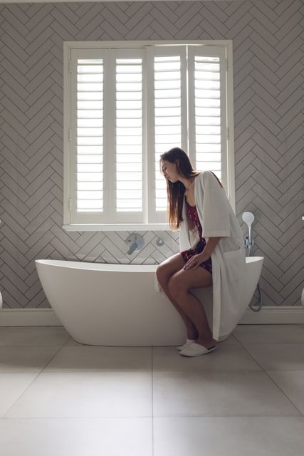 Woman in a white robe checking the water level in a modern bathtub at home. The bathroom features a contemporary design with herringbone tile walls and large windows with shutters. Ideal for use in lifestyle blogs, home decor magazines, and advertisements promoting relaxation and self-care routines.