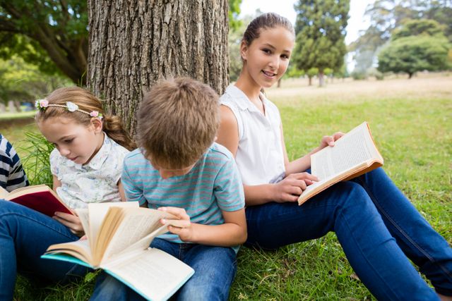 Children sitting under a tree in a park, enjoying reading books on a sunny day. Ideal for educational content, outdoor activities, childhood learning, and leisure time themes. Perfect for use in school brochures, educational websites, and family-oriented advertisements.