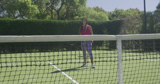 Woman smiling while playing tennis on an outdoor court during a sunny day. Tennis net in foreground with greenery and trees in the background. Ideal for promoting outdoor sports, active lifestyle, and fitness activities.