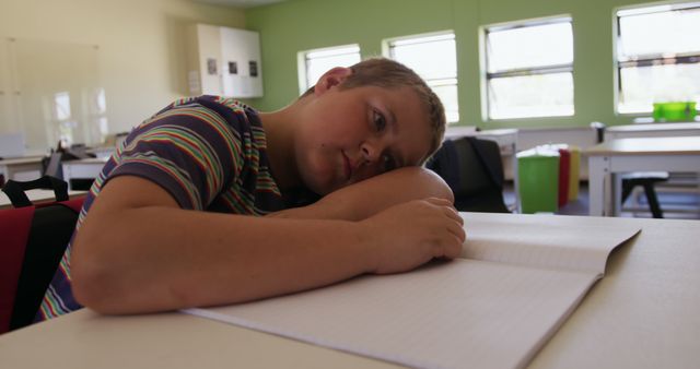 A young student in a striped shirt is resting his head on a desk, looking tired in a brightly lit classroom. The image depicts fatigue and lack of engagement in an educational setting, making it useful for articles or materials related to education, student well-being, academic burnout, or classroom environments.