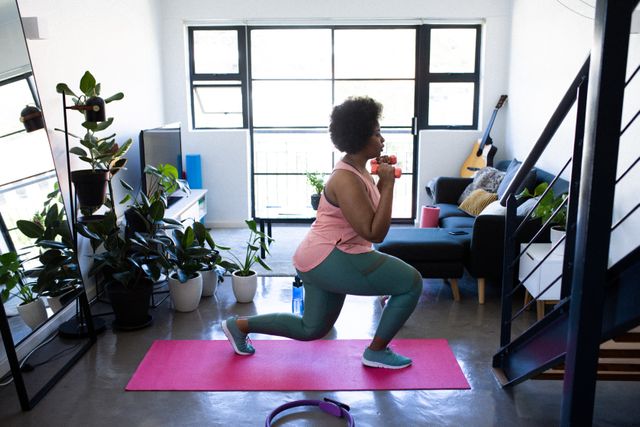 African American woman in sportswear performing lunges with dumbbells in a bright living room. Surrounded by indoor plants and modern furniture, she is focused on her home workout routine. Ideal for content related to home fitness, self-care during isolation, and maintaining a healthy lifestyle indoors.