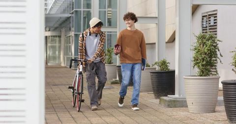 Diverse young men walking college campus pushing bike checking smartphone holding folder