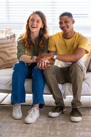 Multiracial couple sitting on sofa holding hands wearing smartwatches sunlit living room