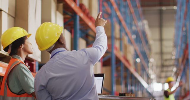 Warehouse workers wearing hard hats and safety gear are inspecting inventory on high shelves. One worker is pointing upwards while explaining something. A laptop is visible, indicating the use of technology for inventory management. Useful for topics on logistics, industrial operations, safety protocols, and teamwork in workplace settings.