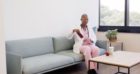 African american physician sitting on sofa checking phone in modern clinic lounge