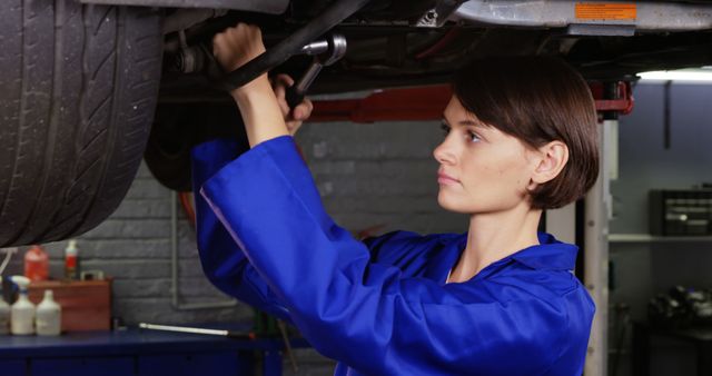 Woman mechanic in blue coveralls fixing car underbody with a wrench in a garage. Ideal for content related to women in STEM, automotive repair services, professional mechanic training, and equal workplace opportunities. Suitable for illustrating professional garage environments and hands-on mechanical skills.