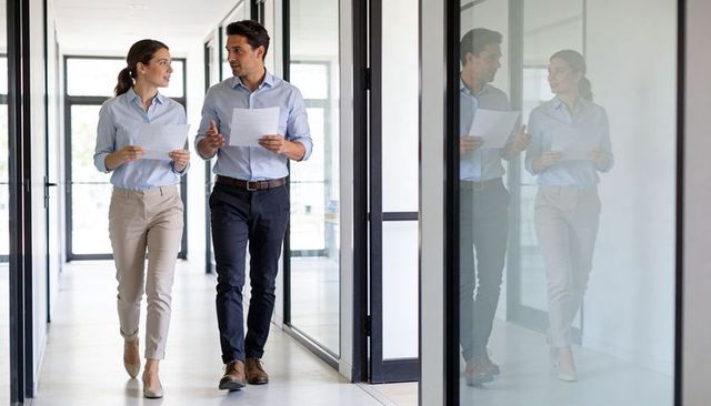 Two coworkers walking and discussing documents in modern glass office corridor