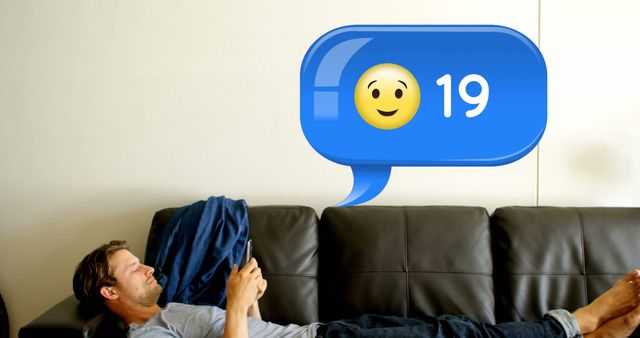 Man lying on couch, using smartphone, receiving emoji notification with number 19. Perfect for content on relaxation, digital communication, social media engagement, modern living, or technology usability studies.