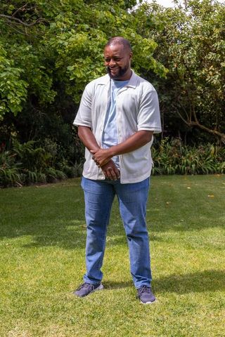 Man standing on sunlit backyard lawn wearing casual button-up shirt and jeans, smiling