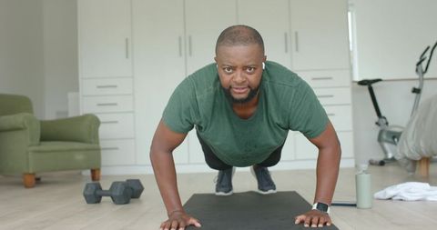 African man doing push-ups during home workout with smartwatch and dumbbells