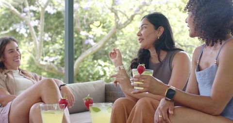 Multicultural friends chatting and sipping cocktails on patio sofa, relaxed summer gathering