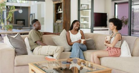 African american friends chatting on sectional couch holding mugs in sunlit living room