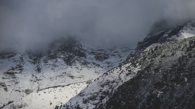 Image captures snow-covered mountain peaks enveloped in mist under an overcast sky. Suitable for use in outdoor adventure promotions, nature photography collections, winter tourism marketing, or wilderness exploration themes. Ideal for illustrating rugged terrains and dramatic weather conditions.