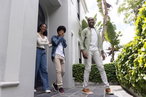 Diverse family greeting on front porch with shy teen covering face, father walking by palms