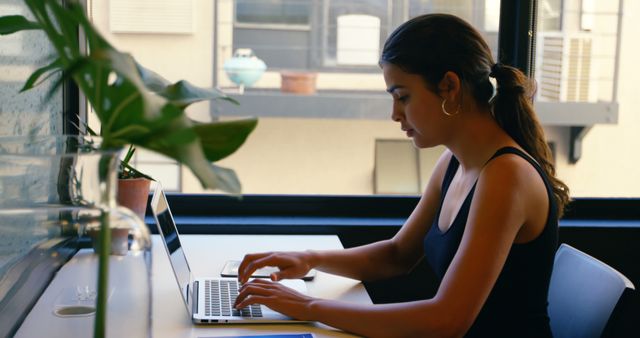 A young woman is focused on her work at a laptop in a home office environment, with copy space. Her professional demeanor suggests she could be engaged in tasks such as remote work, online communication, or research.
