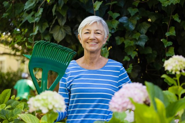 Senior woman enjoying gardening in backyard, holding rake and smiling. Ideal for use in articles or advertisements related to gardening, senior activities, healthy lifestyles, and outdoor hobbies. Perfect for illustrating themes of retirement, nature, and active living.
