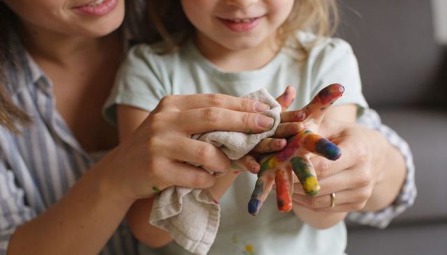 Parent wiping colorful paint from child's hand with soft cloth, home craft bonding moment