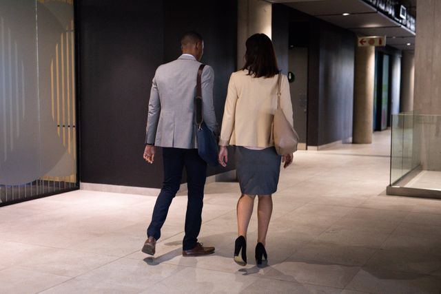 This image depicts a diverse male and female colleague walking through a modern office corridor, carrying bags and engaging in conversation. Ideal for use in business-related content, corporate websites, presentations, and articles focusing on workplace diversity, professional environments, and business communication.