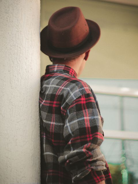 Young man wearing a brown hat and red plaid shirt lounging casually against a light-colored exterior wall. Perfect for fashion lifestyle magazines, urban style blogs, casual clothing advertisements, and metropolitan living promotions.