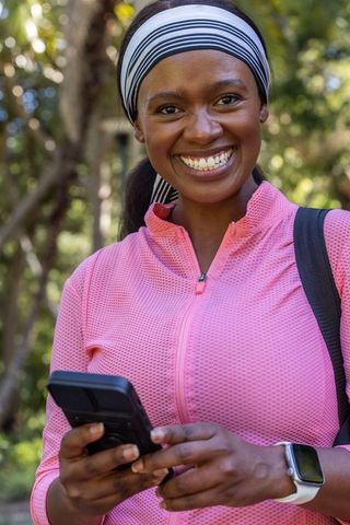 Smiling african woman using smartphone outdoors in pink activewear with smartwatch