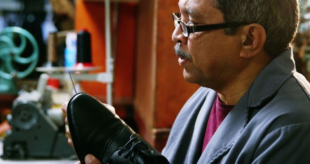 Mature shoemaker wearing glasses works diligently on a black leather shoe in a workshop. He showcases traditional craftsmanship and attention to detail. Surrounded by shoemaking tools, the image highlights the dedication and skill involved in handcrafted shoe repair. Suitable for advertisements regarding traditional crafting skills, shoemaking services, professional artisans, and workshops keeping heritage alive.