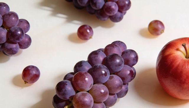 Glossy Purple Grape Cluster with Dew Drops and Red Apple on Cream Surface Closeup