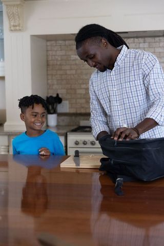 African american dad packing lunch while smiling son watching at kitchen island