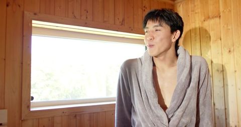 Young asian man relaxing in light gray bathrobe by window inside wood-panel cabin