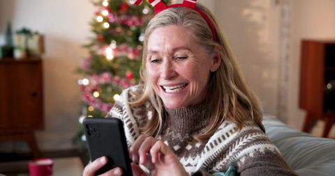 Smiling woman with antler headband using smartphone near christmas tree