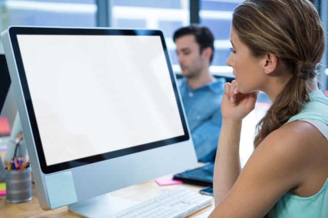 Woman concentrating on desktop computer in a modern office environment. Ideal for illustrating professional work settings, business technology, corporate environments, and productivity themes. Can be used in articles, blogs, and advertisements related to office work, technology in the workplace, and professional development.