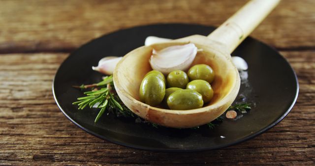 A wooden ladle holds green olives and is placed on a black plate with garlic cloves and rosemary, with copy space. Capturing the essence of Mediterranean cuisine, the image showcases ingredients commonly used in healthy and flavorful dishes.