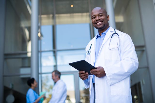 This image shows a smiling doctor holding a digital tablet outside a hospital. The doctor is wearing a white coat and a stethoscope, indicating his profession. In the background, two other medical professionals are engaged in conversation. This image can be used for healthcare-related content, medical technology promotions, hospital advertisements, or articles about modern medicine and healthcare professionals.