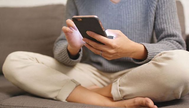 Woman holding and scrolling smartphone on couch legs crossed wearing knit sweater and rolled pants