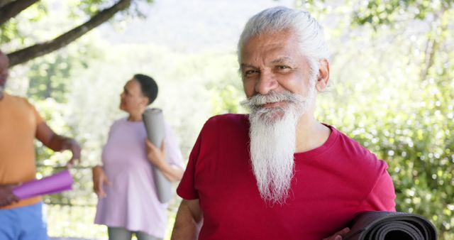 Portrait of happy biracial man with white beard and yoga mat out in sun with friends, slow motion. Summer, friendship, retirement, yoga, wellbeing and healthy senior lifestyle, unaltered.