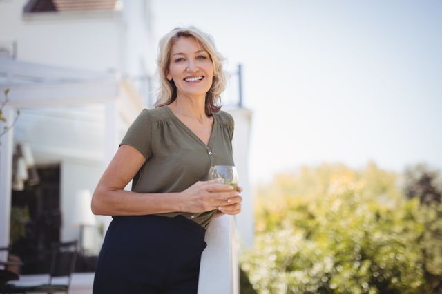 Mature woman smiling and holding a glass of juice outdoors on a sunny day. She is casually dressed and appears relaxed and happy. This image can be used for lifestyle blogs, health and wellness articles, advertisements for home or garden products, and promotional materials for healthy living.