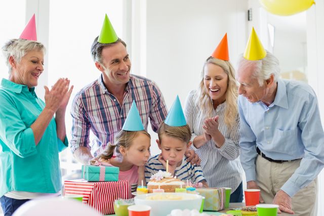 Family members of different generations celebrating a birthday at home. Children and adults wearing party hats, gathered around a table with a birthday cake and presents. Ideal for use in advertisements, family-oriented content, and articles about family celebrations and togetherness.