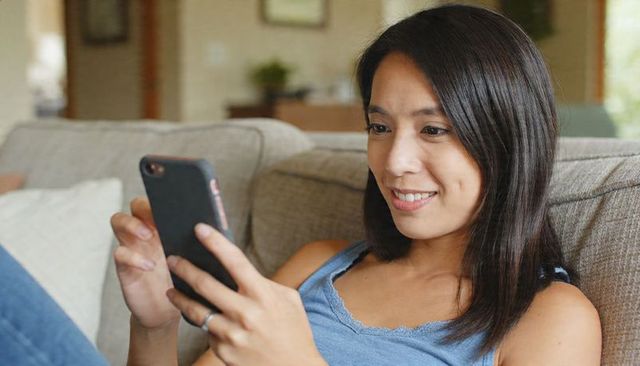 Relaxing on couch using smartphone, smiling woman in casual tank and jeans at home