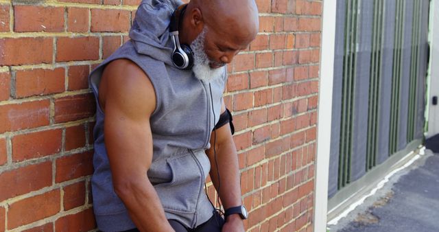 An African American middle-aged man rests against a brick wall during a workout break, with copy space. He appears focused and choosing music or tracking his fitness progress on a wearable device.