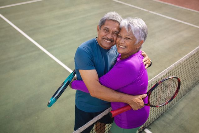 Portrait of smiling biracial senior couple with rackets embracing while standing at tennis court. high angle view, romance, tennis, love, togetherness, unaltered, lifestyle, sport and retirement.