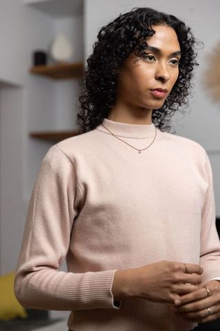 Poised non-binary adult in pale pink sweater wearing gold pendant, minimalist portrait