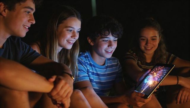 Teen friends watching fireworks on tablet at night outdoors smiling and bonding