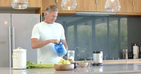 Senior man preparing healthy green smoothie with blender on modern kitchen island
