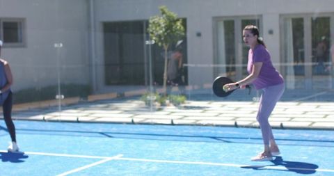 Woman serving pickleball on bright blue outdoor court in purple activewear with glass backdrop