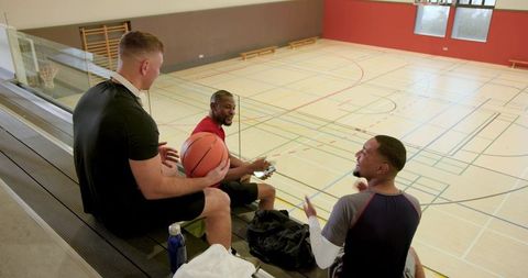 Multicultural male friends spinning basketball and using smartphone on indoor gym bleachers