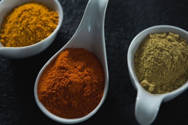 This image shows a close-up view of various spices, including turmeric, paprika, and cumin, placed in white ceramic spoons on a dark background. It is ideal for use in culinary blogs, recipe websites, cooking magazines, and food-related marketing materials to highlight the use of natural and aromatic ingredients in cooking.