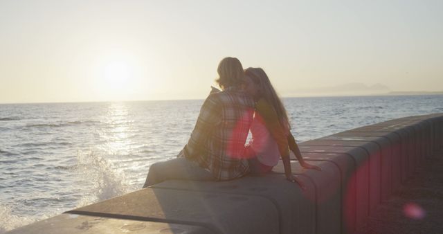 Couple sitting together on a seawall, embracing and enjoying a serene sunset over the ocean. Great for romantic themes, travel destinations, love and relationship concepts, and waterfront real estate promotions.